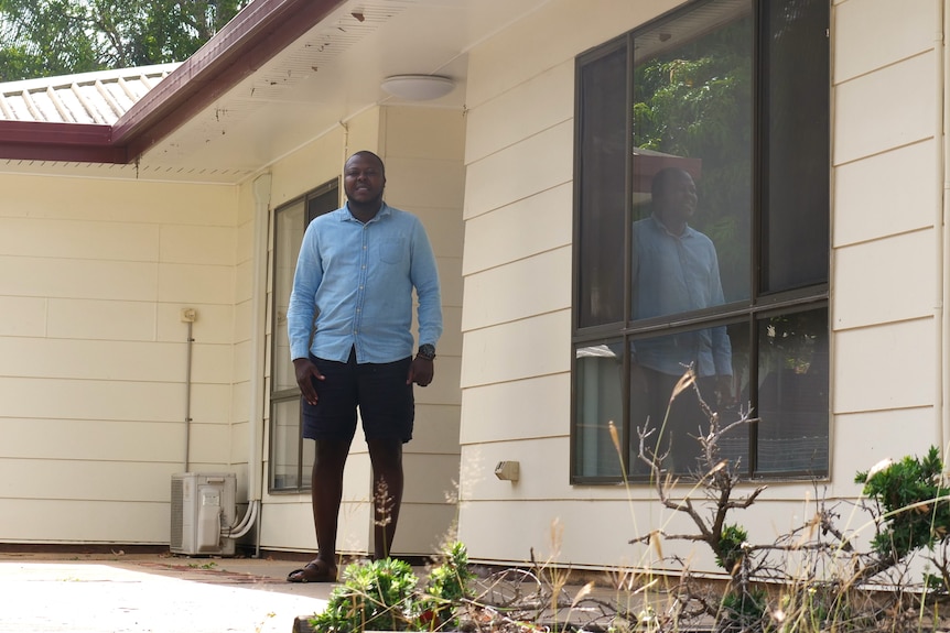 a zamian man stands outside the front of his rural home in black shorts and a blue button up shirt