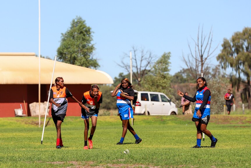 Young Aboriginal women laugh while playing football in brightly coloured guernseys on a green grassed oval