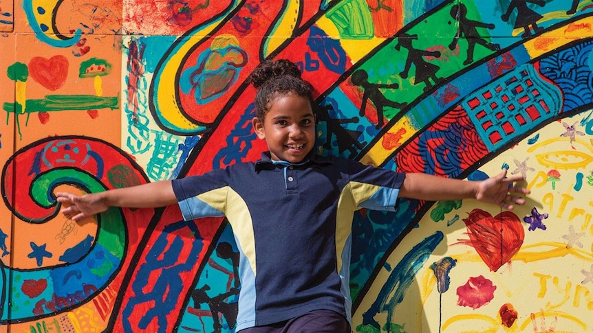 Aboriginal or Torres Strait Islander girl in navy, blue and yellow uniform standing against brightly painted wall mural.