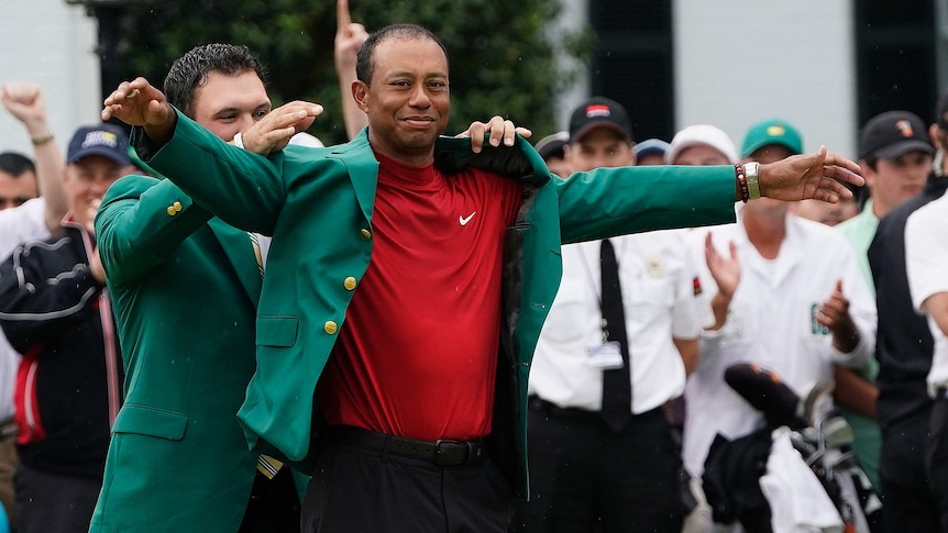 A man helps put a green jacket on another golfer as people applaud.