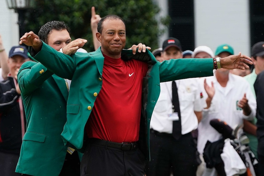 A man helps put a green jacket on another golfer as people applaud.