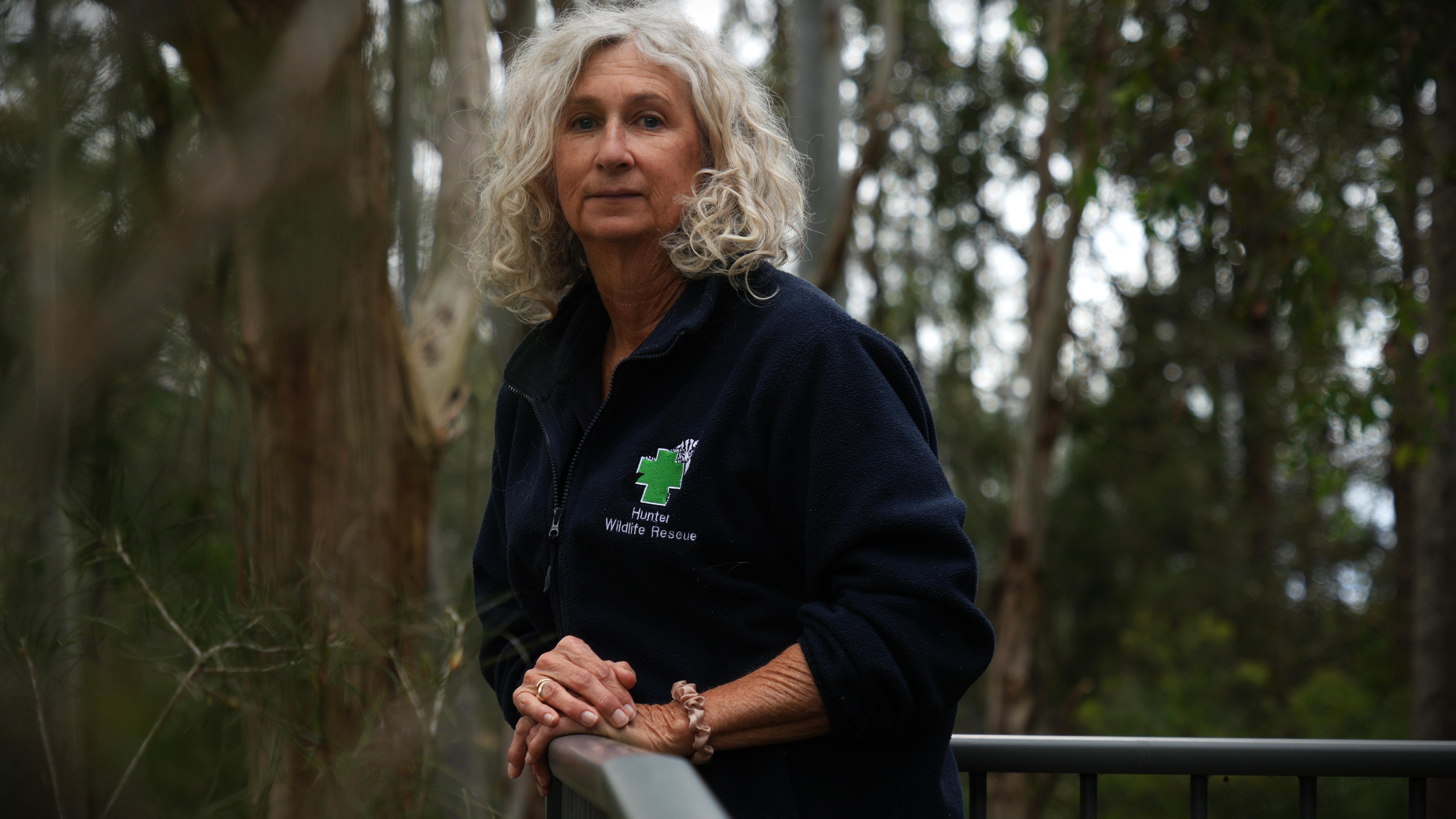 Kerry Walker has short hair, wears zip up jacket and leans on a fence overlooking a forest. 