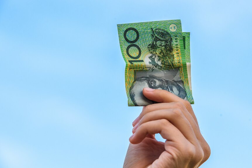 A woman's hand holding a $100 note in the air, against a blue sky.