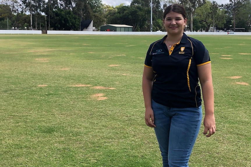 Alyssa Kelly wearing a dark polo shirt and blue jeans, cricket field, grass behind.