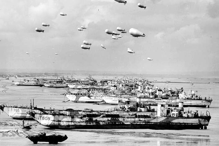 Black and white photo of British ships lining the Normandy shore each with a barrage balloon attached.
