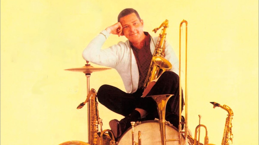 Arranger Bill Holman in a white jacket, sitting on a bass drum surrounded by his big band's instruments
