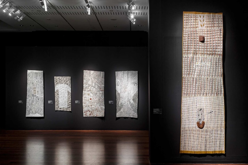 Darkened gallery with black walls hung with long bark paintings from Arnhem Land.