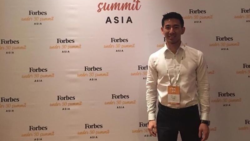 Daniel Liang wearing a white shirt and black suit pants and standing in front of a Forbes sign