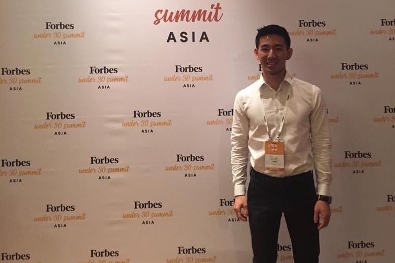 Daniel Liang wearing a white shirt and black suit pants and standing in front of a Forbes sign