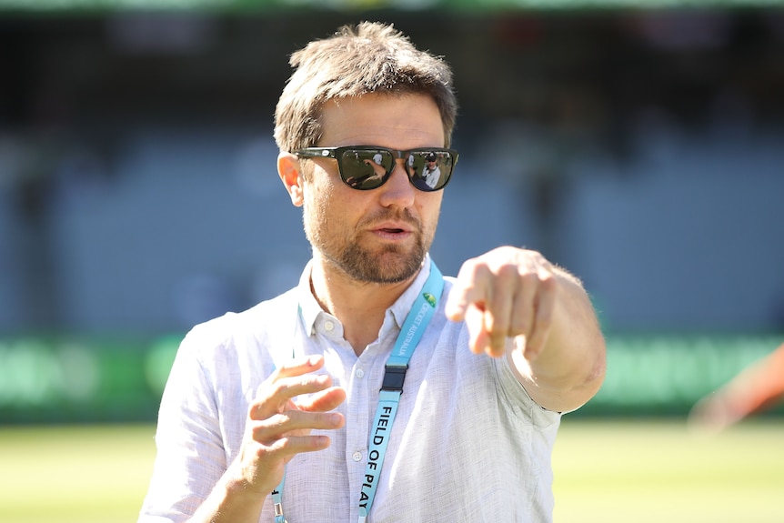 Dirk Nannes pointe devant lui alors qu'il se tient sur un terrain de cricket avec un polo et des lunettes de soleil.