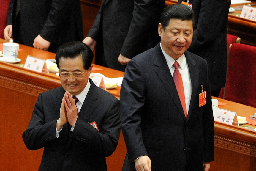 Younger Hu Jintao with black hair and hands pressed together as Xi walks past glancing at him.