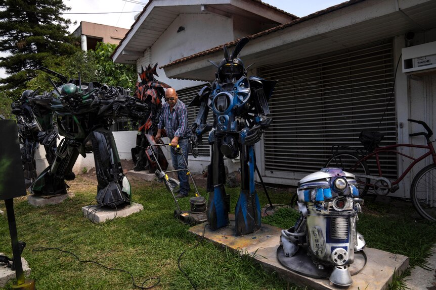 A man stands on grass with a lawnmower between transformer statues. 