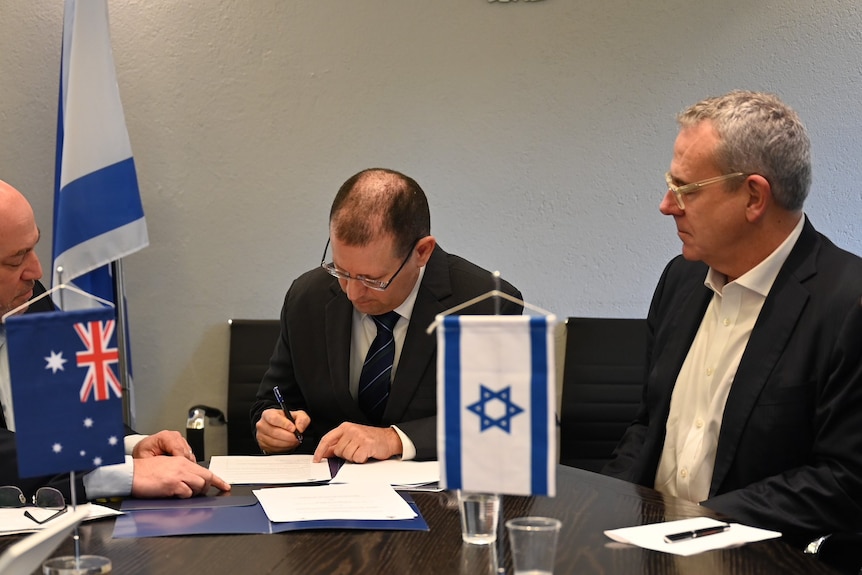 Three middle-aged men in suits sign a document at a desk bearing the Australian and Israeli flags.