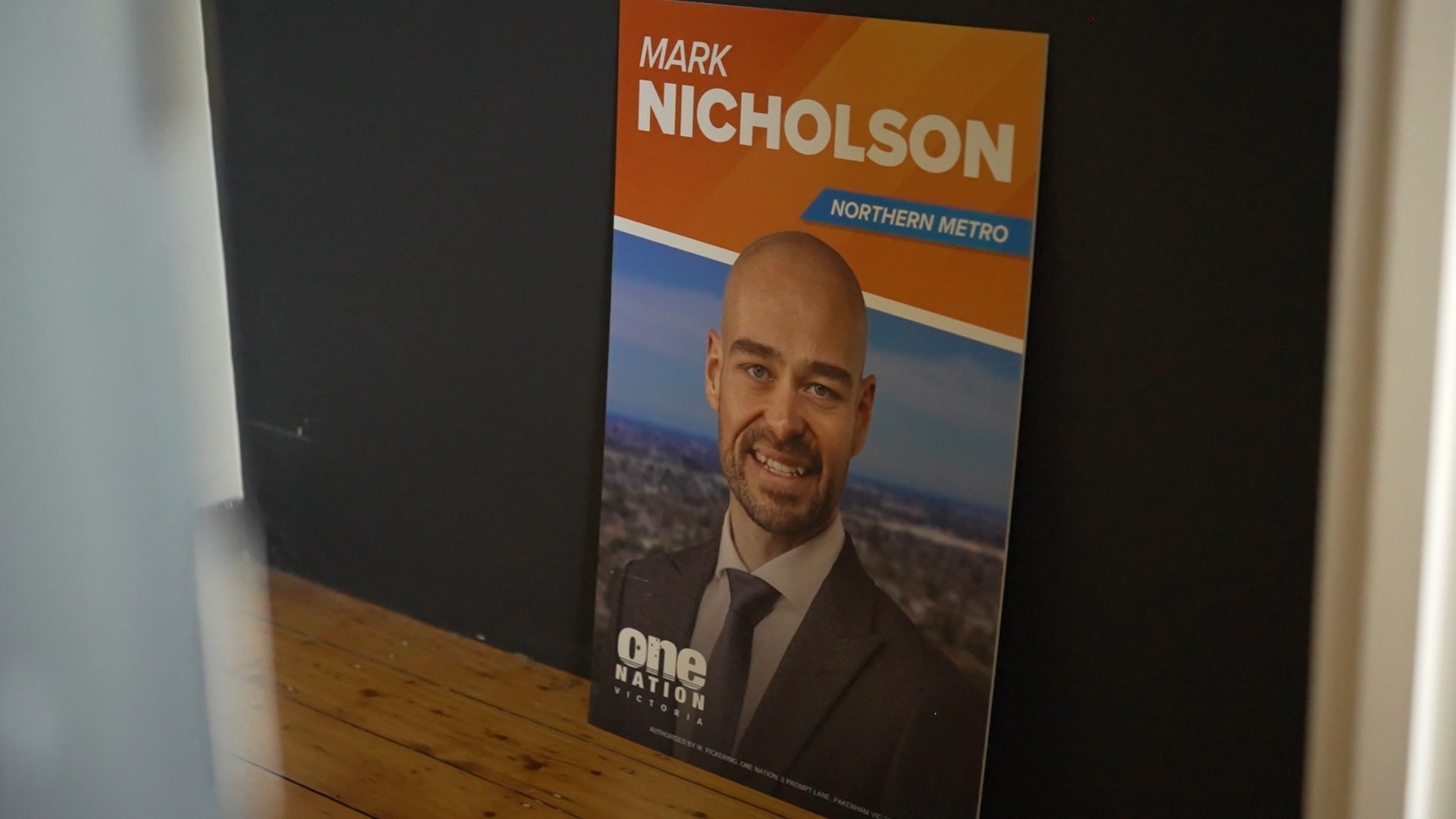 A One Nation corflute for Mark Nicholson sits on a wooden floor leaning against against a dark wall.