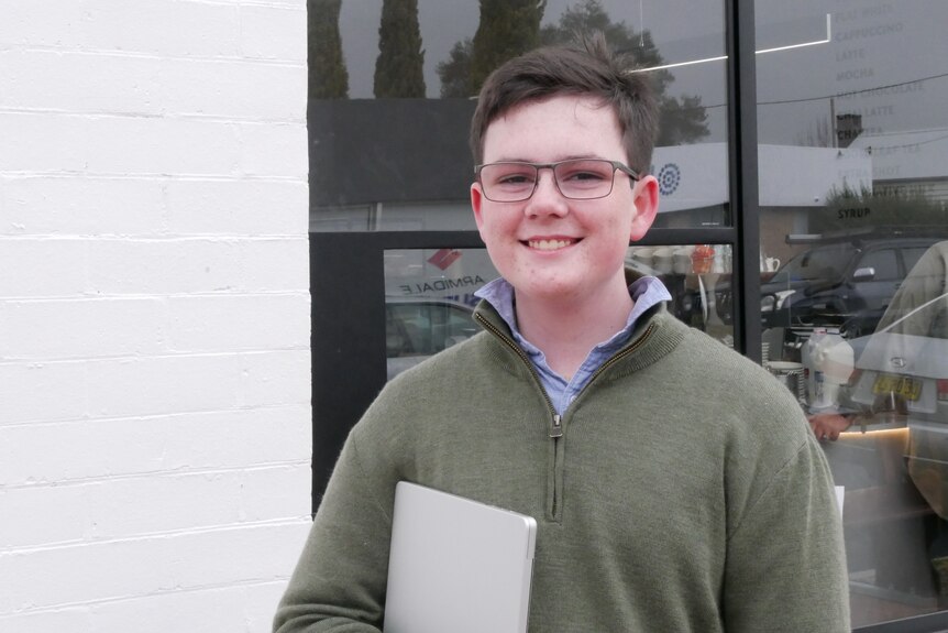 Teenage boy stands out the front of a cafe building, smiling and holding a closed laptop