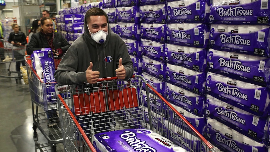 A man in a mask gives a thumb's up as he pushes a shopping trolley with a big pack of toilet paper in it.