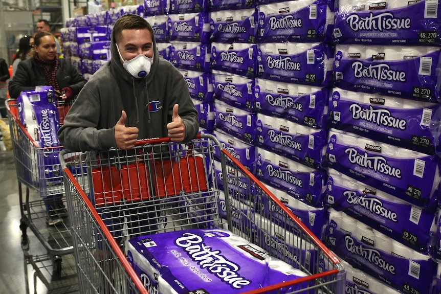 A man in a mask gives a thumb's up as he pushes a shopping trolley with a big pack of toilet paper in it.