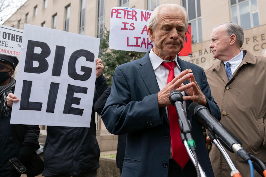 A close up of a man in a suit and tie with his hands together outside a court house. A sign held next to him reads 'BIG LIE'