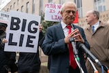 A close up of a man in a suit and tie with his hands together outside a court house. A sign held next to him reads 'BIG LIE'