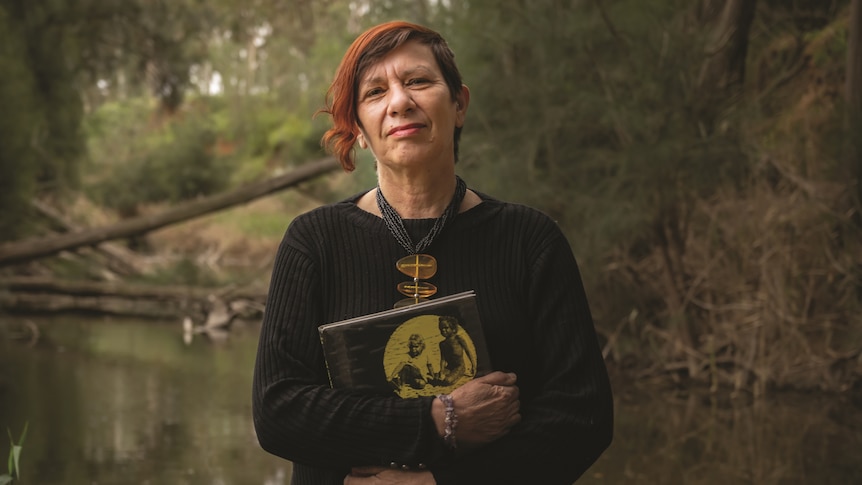 An Aboriginal woman in her late 50s stands in front of a swamp holding a book