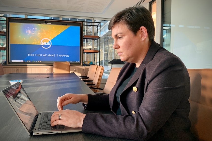 A woman with cropped dark hair wearing a merlot-coloured blazer sits at a conference table in a board room working on a laptop.