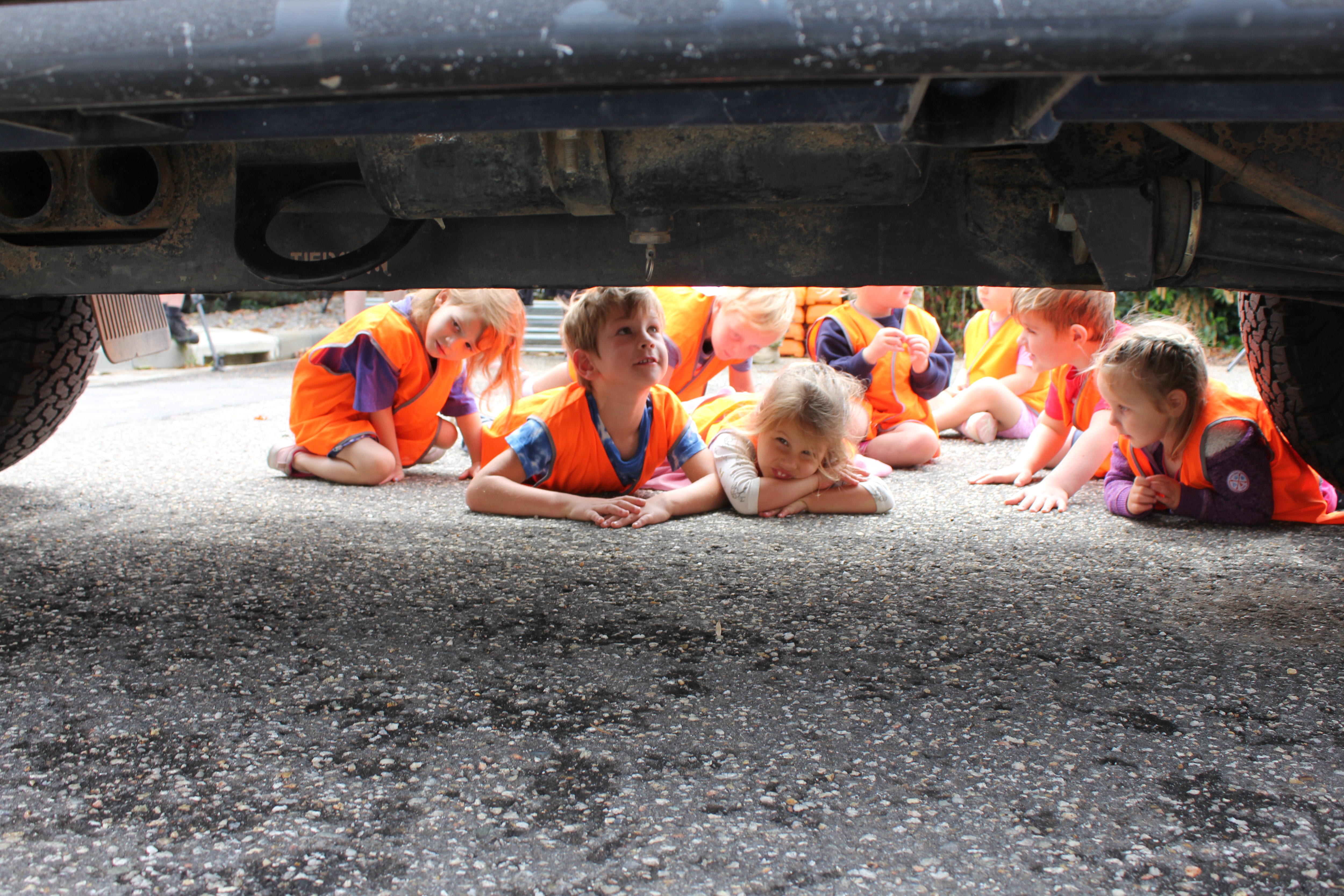 A group of very young children, wearing high-vis vests, peer under the underside of an army vehicle.