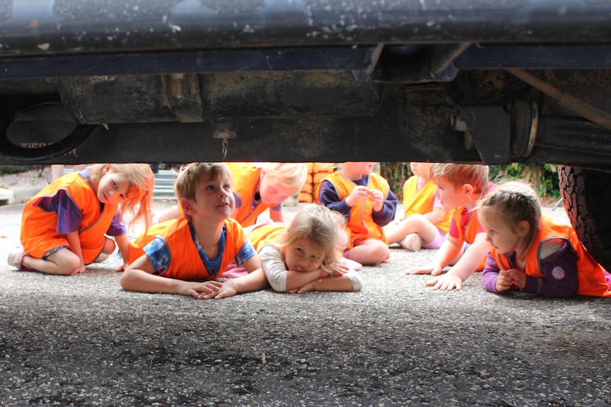 A group of very young children, wearing high-vis vests, peer under the underside of an army vehicle.