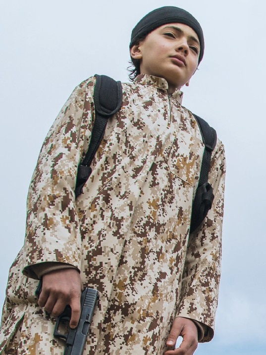 A boy holds a hand gun while wearing military uniform and an Islamic headband.