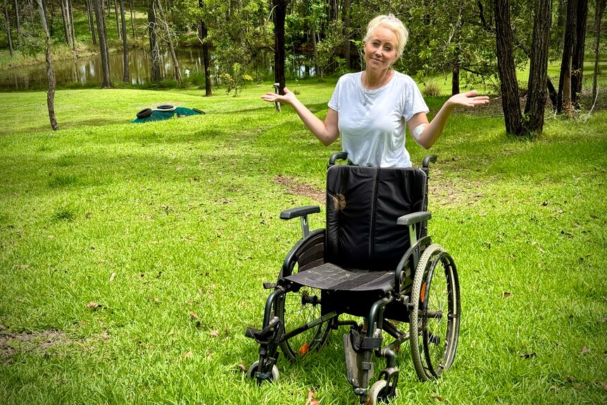 A woman holds up her hands as she stands behind an empty wheelchair on a steep hill with a dam at the bottom.