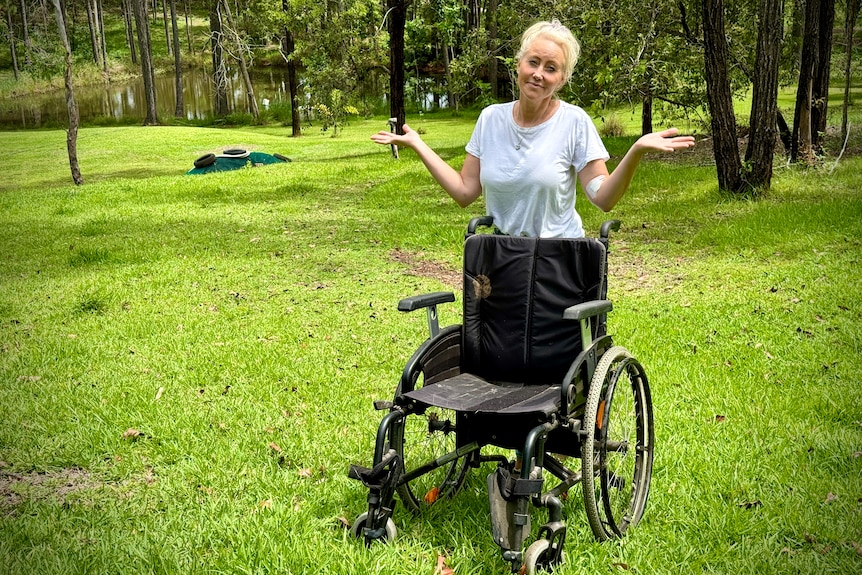 A woman holds up her hands as she stands behind an empty wheelchair on a steep hill with a dam at the bottom.