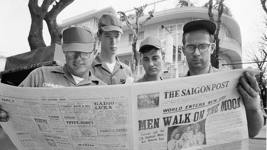 Four men in uniform pore over a newspaper covering the Apollo 11 mission