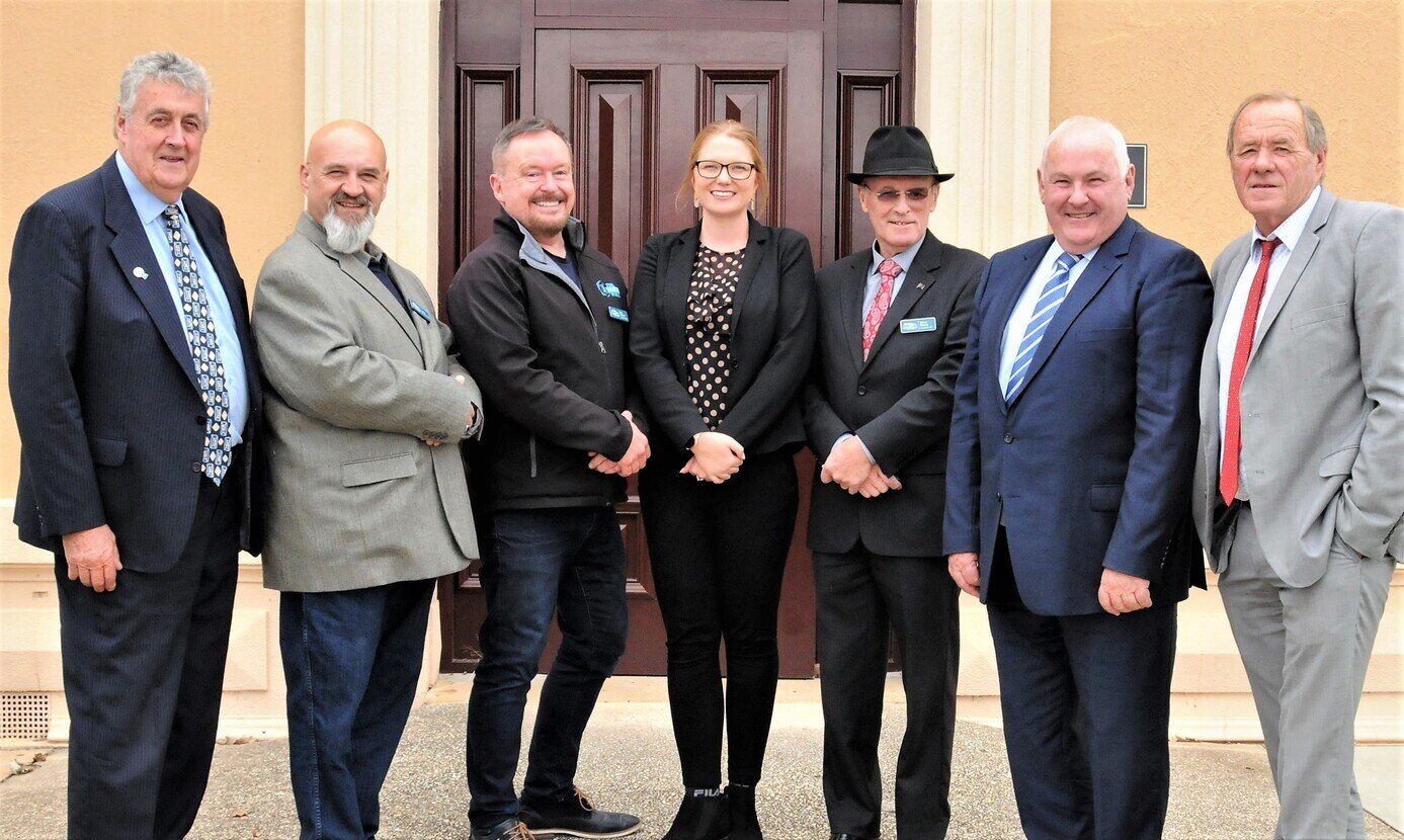 A group photo of six men and one woman in business attire in front of a large wooden door and orange walls.  All are smiling.