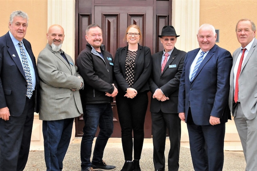 A group photo of six men and one woman in business attire in front of a large wooden door and orange walls.  All are smiling.