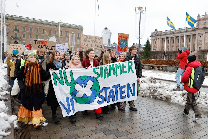 a group of young activists hold up a sign which reads "now we sue the state" as they walk through Stockholm
