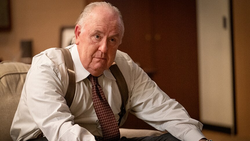 An elderly man in suspenders, button up shirt and tie sits on couch in office with serious expression.