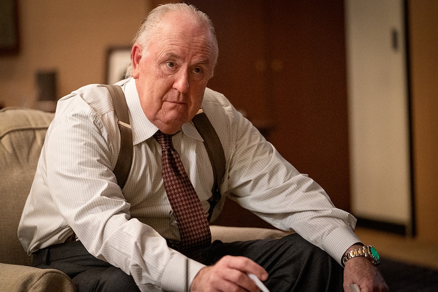 An elderly man in suspenders, button up shirt and tie sits on couch in office with serious expression.