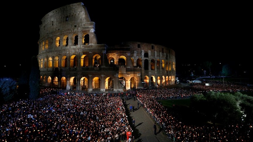 A large crowd holding candles gathers in the dark outside Rome's Colosseum