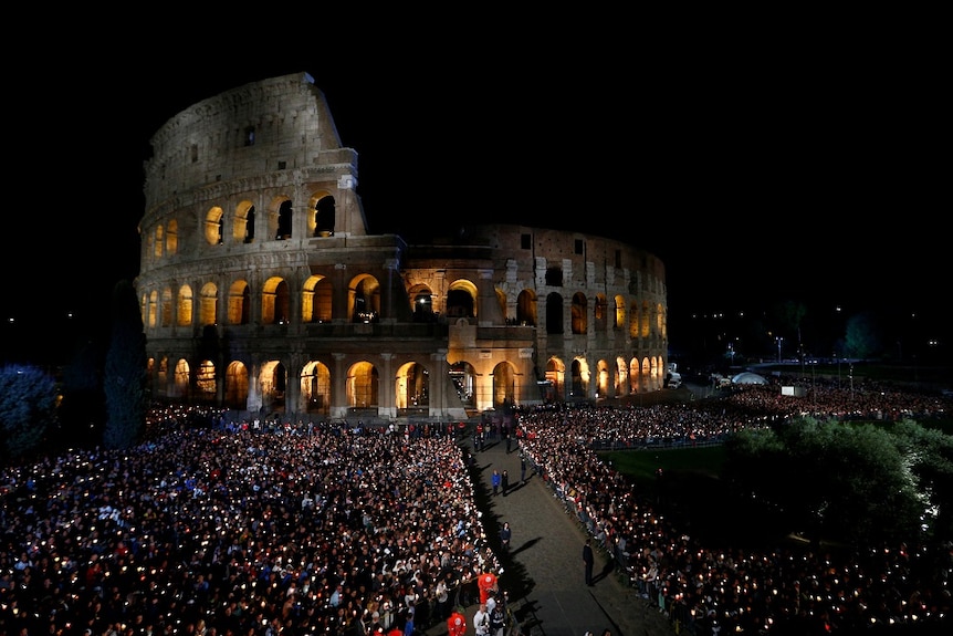 A large crowd holding candles gathers in the dark outside Rome's Colosseum
