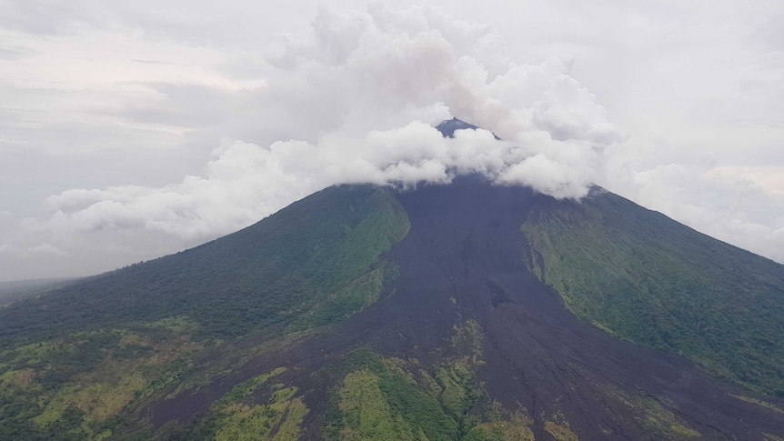 Smoke rises from a volcanic mountain and black lava can be seen on the mountainside.