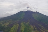 Smoke rises from a volcanic mountain and black lava can be seen on the mountainside.