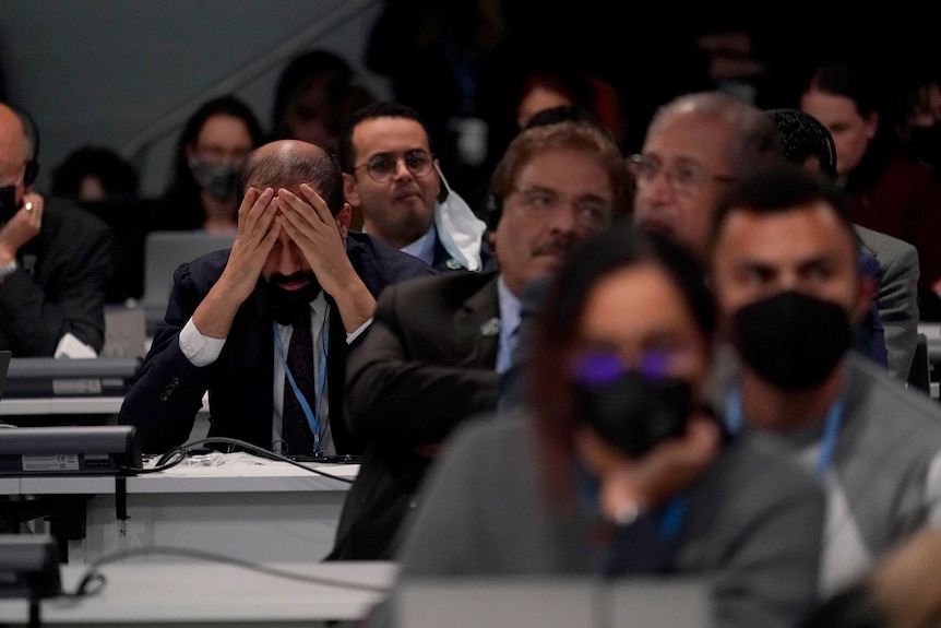 A man sits with his head in his hands among others in a conference room.