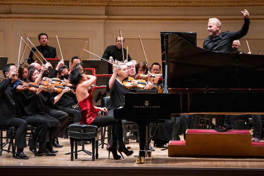Yuja Wang plays a grand piano in front of an orchestra in a sparky red dress. Her right arm is thrown up dramatically.