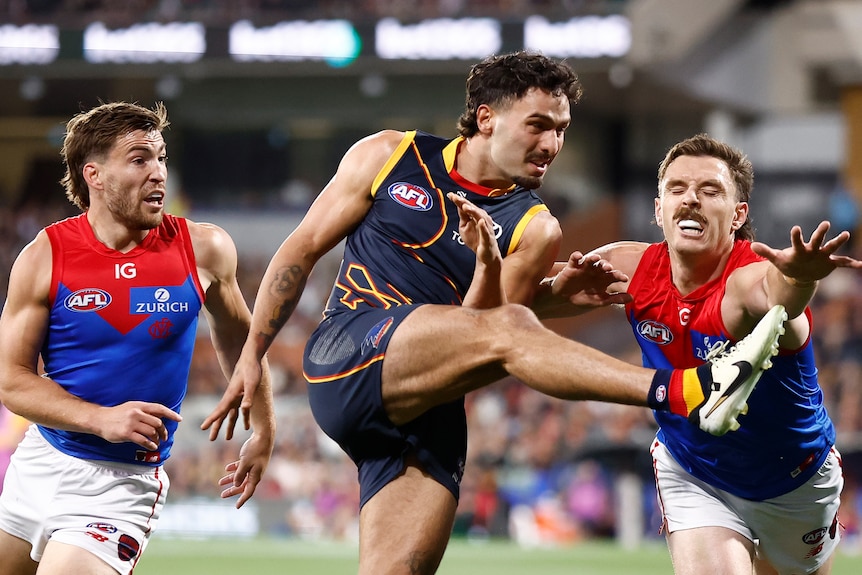An Adelaide Crows AFL player kicks the ball as a Melbourne defender extends his hand out trying to block the kick.