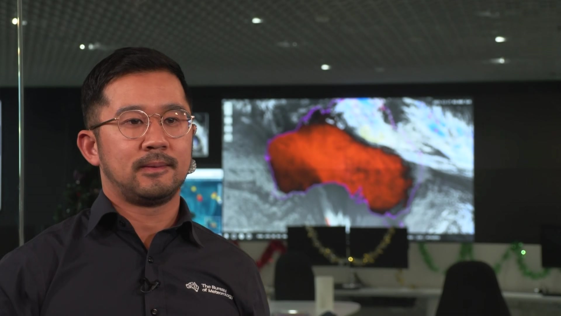 Un hombre con cabello oscuro y gafas, vestido con una camisa de cuello negro, se para frente a una pantalla que muestra un gráfico de Australia.