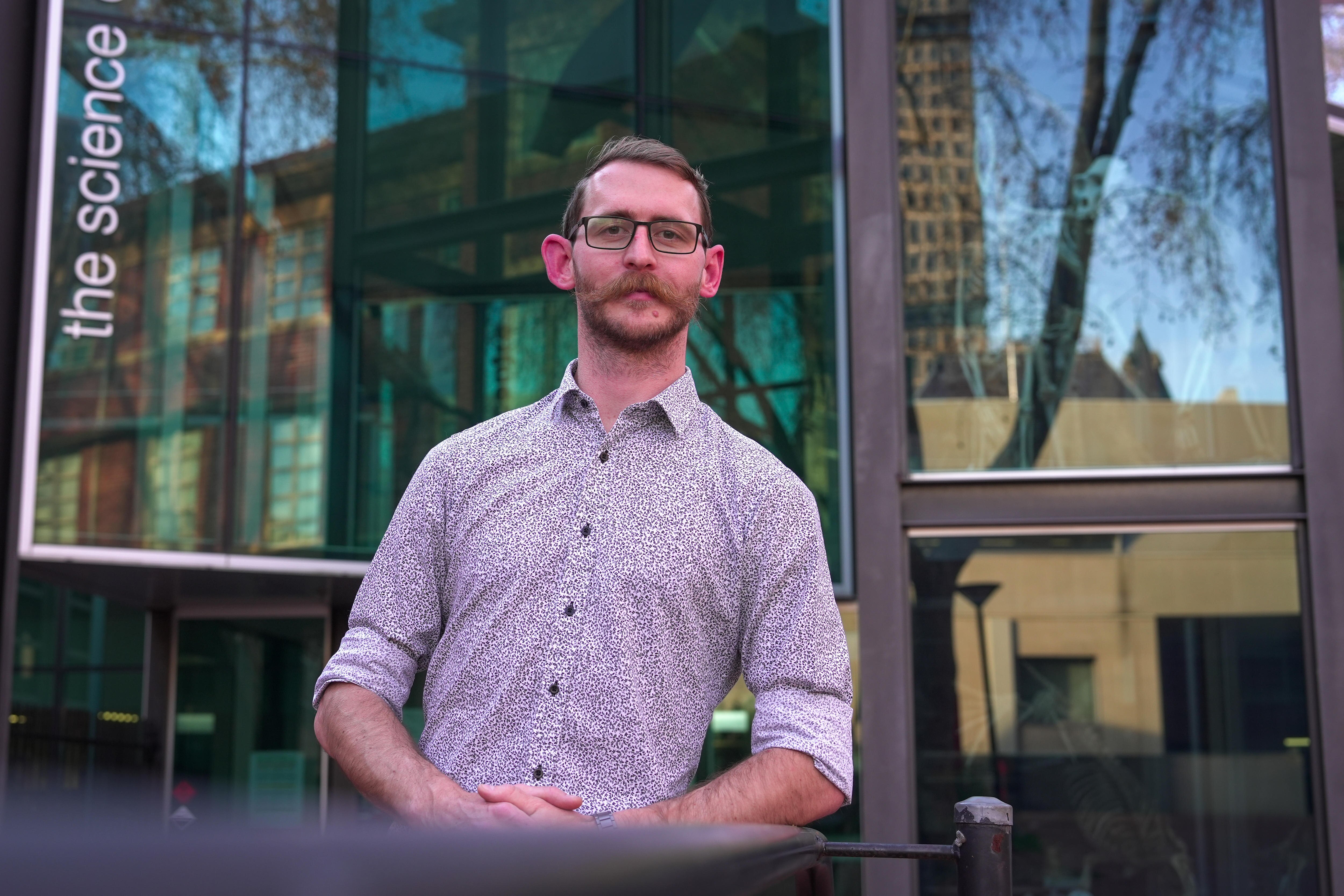 A man with short hair, a beard and large moustaches stands outside a building with a glass facade.