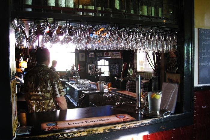 A dark bar in a pub can be seen, a bartender has his back to camera, with empty glasses and lots of bottles in picture.