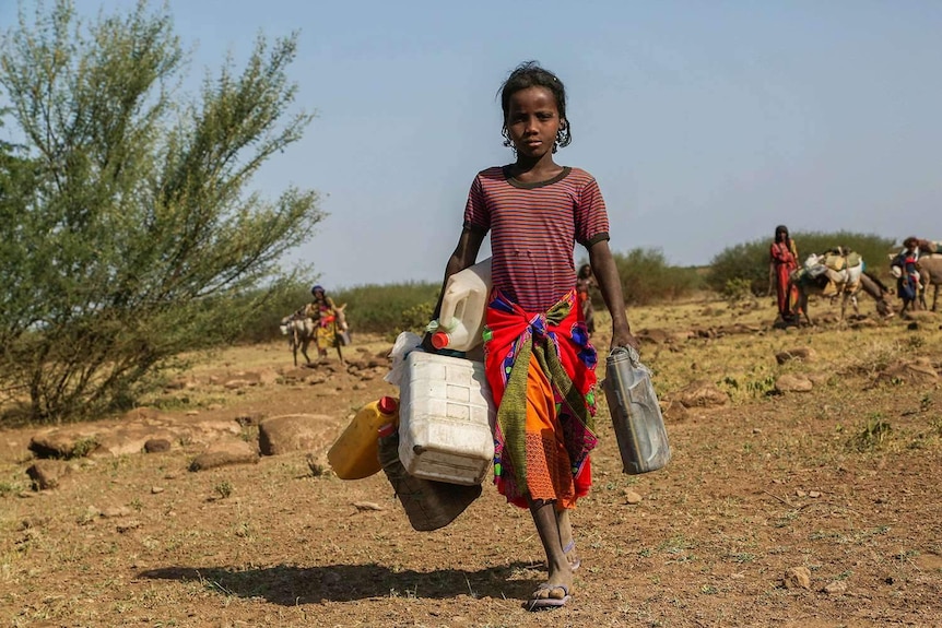 A girl carrying several water containers against an agricultural background.