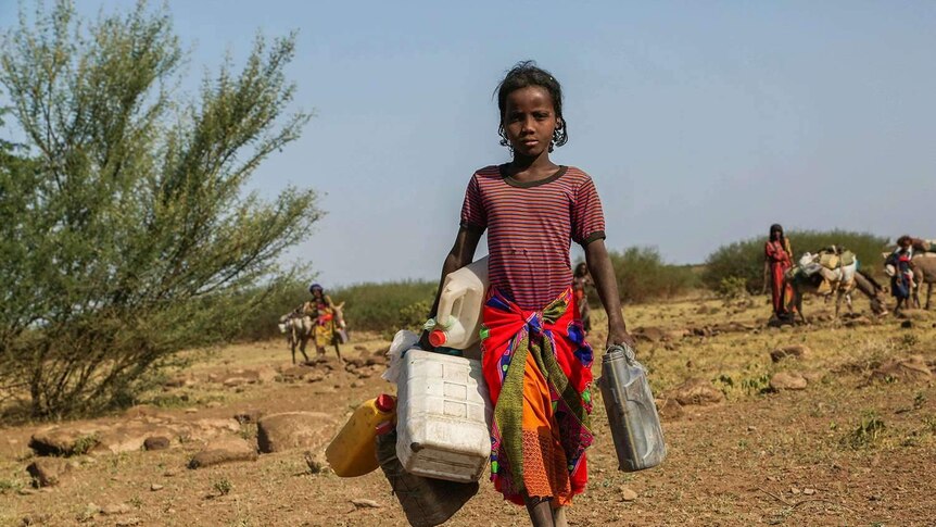 A girl carrying several water containers against an agricultural background.
