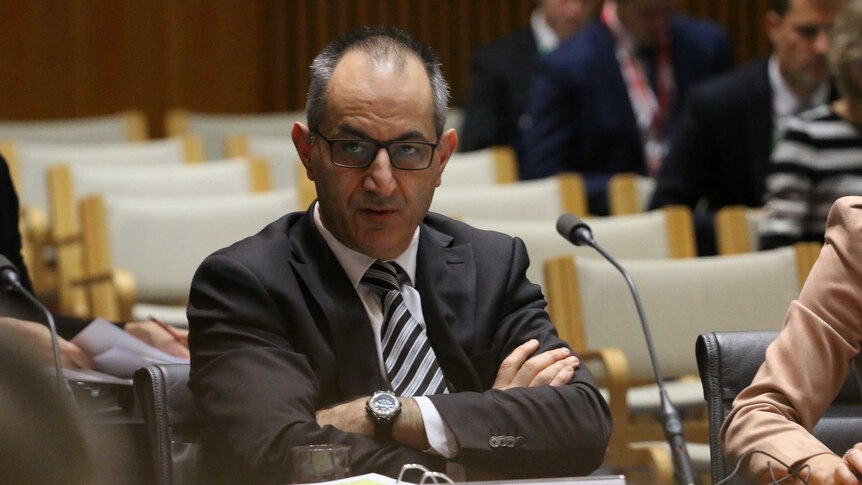 Mike Pezzullo wears black framed spectacles, a brown suit and striped tie as he crosses his arms in a senate estimates hearing.