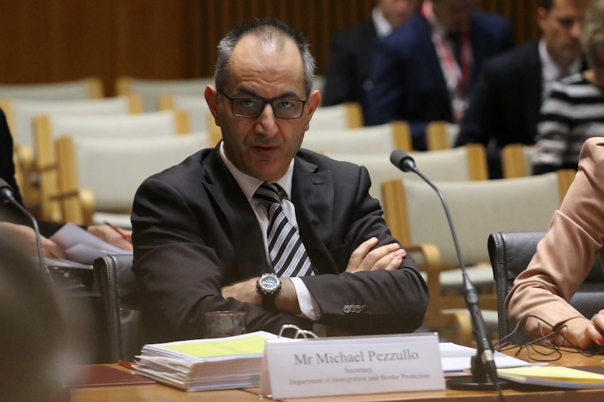 Mike Pezzullo wears black framed spectacles, a brown suit and striped tie as he crosses his arms in a senate estimates hearing.